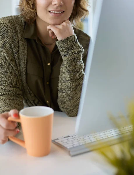 Attraktiv leende kvinna sitter vid skrivbordet, håller en kopp kaffe, hon är avkopplande och tittar bort — Stockfoto