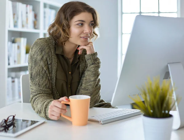 Mulher sorridente atraente sentado na mesa do escritório, segurando uma xícara de café, ela está relaxando e olhando para longe — Fotografia de Stock