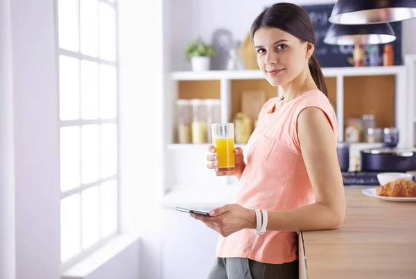 Leende vackra kvinna tittar på mobiltelefon och hålla glas apelsinjuice medan du äter frukost i ett kök. — Stockfoto