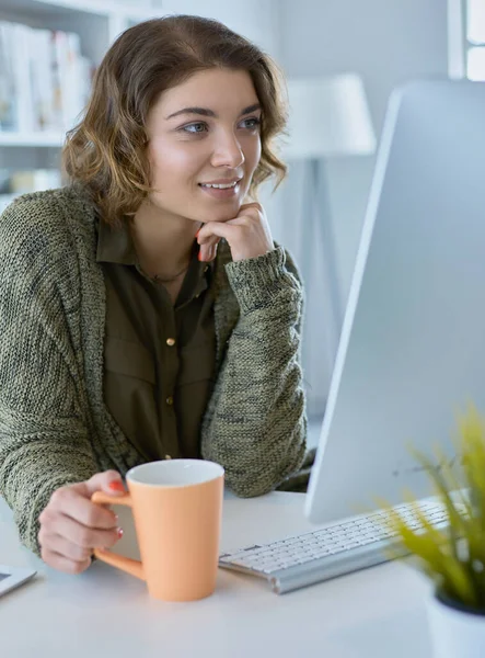 Attraktiv leende kvinna sitter vid skrivbordet, håller en kopp kaffe, hon är avkopplande och tittar bort — Stockfoto
