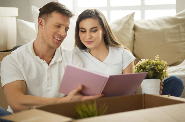 Casal bonito desempacotar caixas de papelão em sua nova casa, sentado no chão e olhando para um álbum de família — Fotografia de Stock