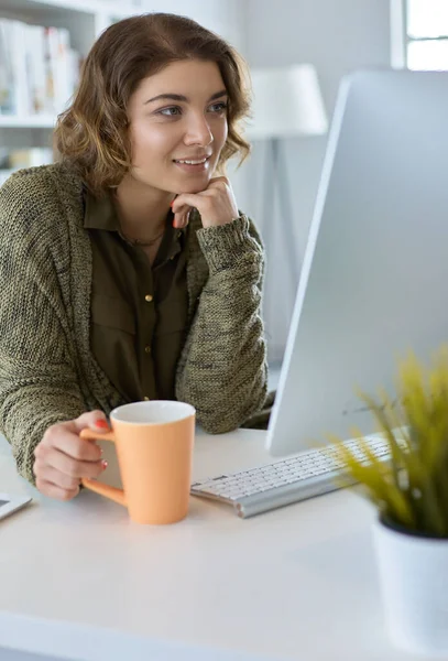 Mulher sorridente atraente sentado na mesa do escritório, segurando uma xícara de café, ela está relaxando e olhando para longe — Fotografia de Stock