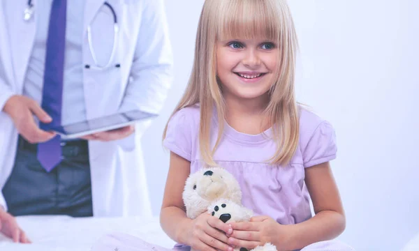 Klein meisje met teddybeer kijkt naar de camera. Vrouwelijke arts op de achtergrond — Stockfoto
