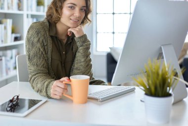 Çekici, gülümseyen bir kadın büro masasında oturuyor, elinde bir fincan kahve tutuyor, rahatlıyor ve başka tarafa bakıyor.