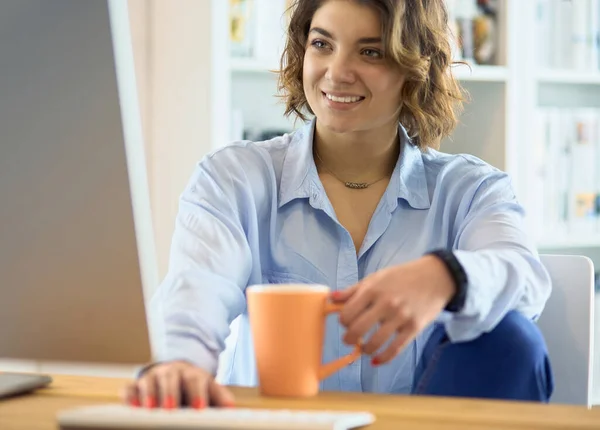 Attraktiv kvinna som sitter vid skrivbordet och håller en kopp kaffe — Stockfoto