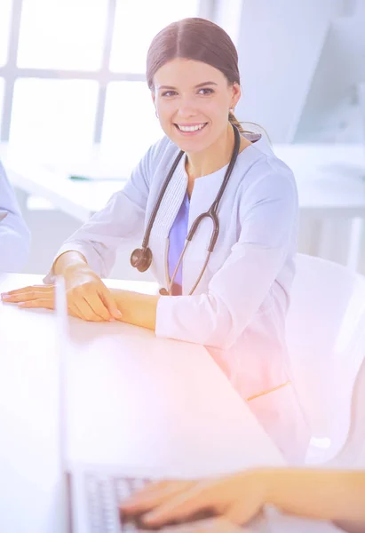 En smilende lege som bruker en bærbar PC til å jobbe med kollegene på et lysende sykehusrom. – stockfoto