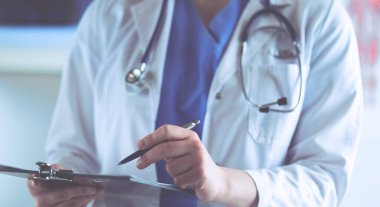 Steteskoplu bir doktor elinde bir defter tutuyor. Hastanede ayakta dikilirken panoda tıbbi form dolduran bir kadın doktorun yakın çekimi.