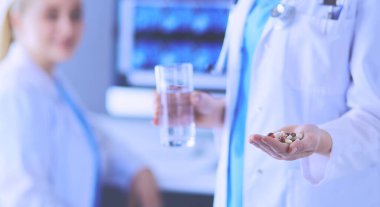Doktorun arka planda meslektaşıyla klinikte elinde haplar ve bir bardak su tutarken görüntüsü..