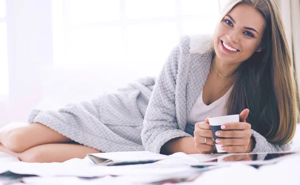 Relaxed young woman sitting on bed with a cup of coffee and digital tablet — Stock Photo, Image