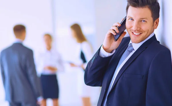 Retrato de jovem empresário no cargo com colegas em segundo plano e usando celular — Fotografia de Stock