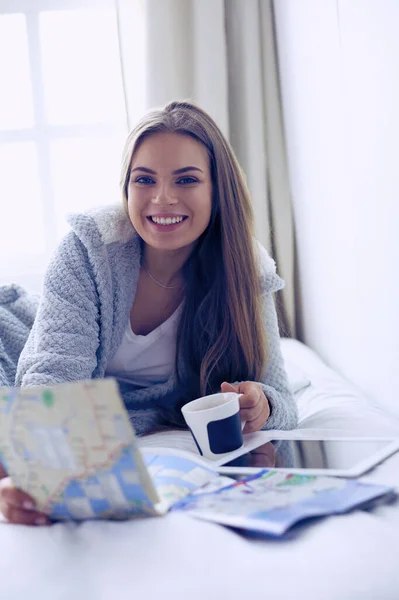 Avslappnad ung kvinna sitter på sängen med en kopp kaffe och digital tablett — Stockfoto