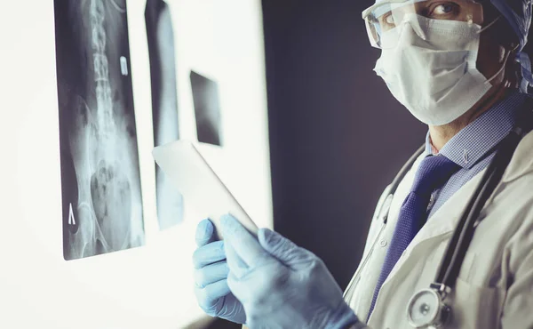 Arzt im Krankenhaus sitzt am Schreibtisch und betrachtet Röntgenbilder auf Tablet vor weißem Hintergrund mit Röntgenaufnahmen — Stockfoto