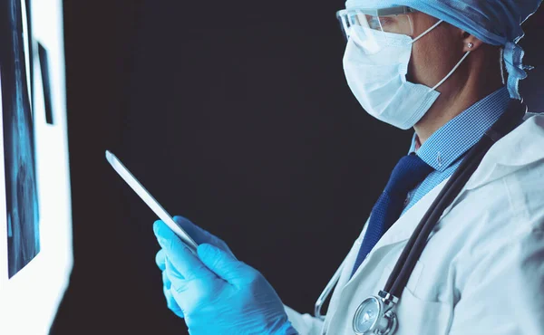 Médico en el hospital sentado en el escritorio mirando los rayos X en la tableta contra el fondo blanco con rayos X — Foto de Stock
