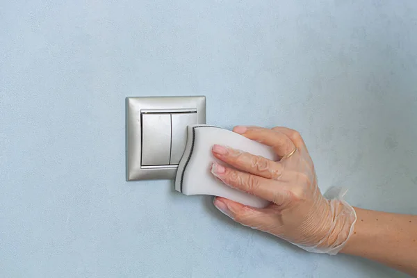 Hand in vinyl gloves is cleaning the light switch on blue wall by melamine sponge.