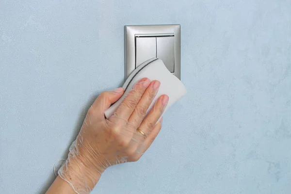 Woman hand in vinyl glove is cleaning the light switch on blue wall by melamine sponge.
