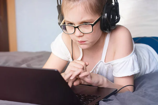 Smart girl in eyeglasses and headphones looking at the screen and doing lessons on laptop. E-learning concept