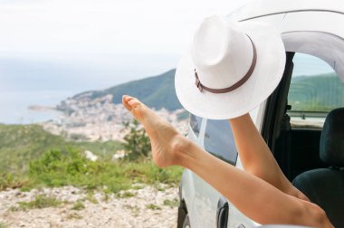 Kadın bacaklar bir gökyüzü, deniz ve dağ manzaralı bir araba pencereden dışarı asılı. Kadın rahatlatıcı bir yolculuğa bir arabada