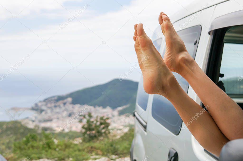 Female feet hanging out of car window at sunset. Woman relaxing in a car on road trip