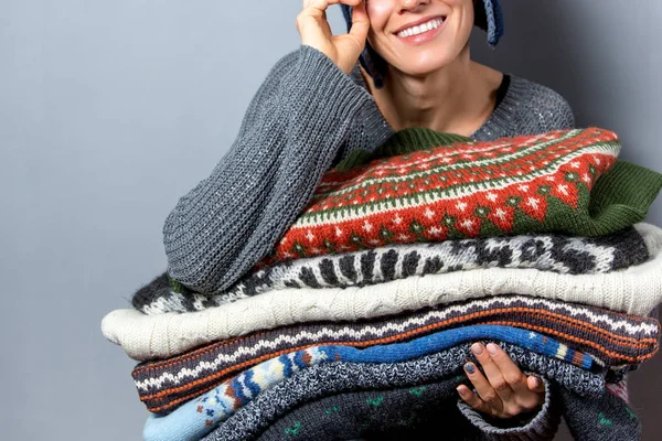 Young woman hold pile of sweaters wearing knitted hat and warm wool sweater