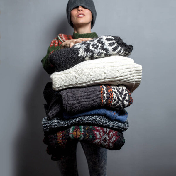 Young woman hold pile of sweaters wearing knitted hat and warm wool sweater, focus on clothes