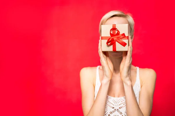 Young happy woman with a gift covering her face by a gift while standing against red background