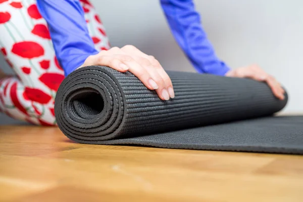 Time for meditation. Girl rolling fitness mat before, after class at home on wooden floor. Hands and legs close up, focus on mat