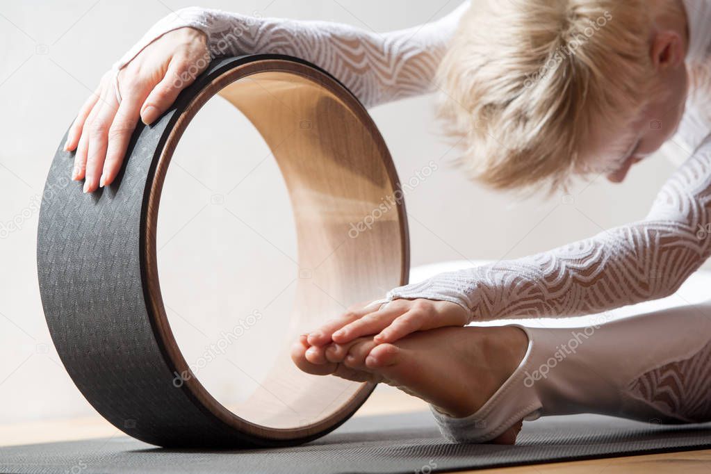 Portrait of gorgeous young woman practicing yoga indoor with a help of yoga wheel.