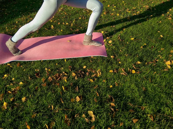 Yoga girl in autumn park with yoga mat on green carpet with fallen yellow leaves. Funny autumn legs wearing woolen socks and doing yoga in cold autumn park in the morning