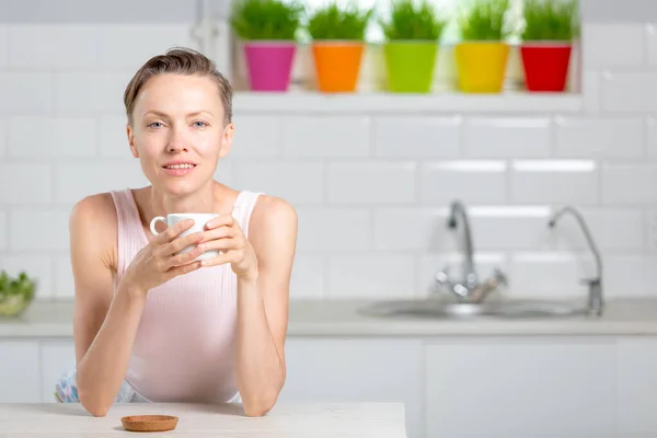 Balance in life concept. Beautiful woman drinking tea in white kitchen with green wheatgrass for juice on the background