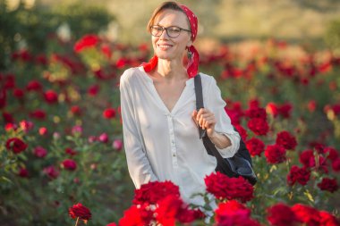 Kırmızı başörtüsü takan güzel bir kadın, beyaz bluz kırmızı gül tarlasında duruyor.