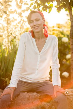 Doğal makyajlı mutlu genç bir kadın beyaz bluz giyiyor, kafasında kırmızı eşarp ve gri pantolon var, doğal ve neşeli genç bir kadın dinleniyor ve güneşli günün tadını çıkarıyor.