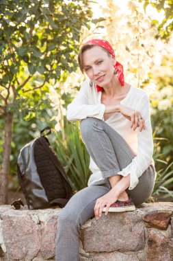 Doğal makyajlı mutlu genç bir kadın beyaz bluz giyiyor, kafasında kırmızı eşarp ve gri pantolon var, doğal ve neşeli genç bir kadın dinleniyor ve güneşli günün tadını çıkarıyor.