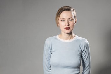 Kısa saçlı, açık gri arka planda doğal makyajı olan genç bir kadın. Spor tişörtlü ve spor formalı güzel sarışın bir kadının kapalı bir portresi.