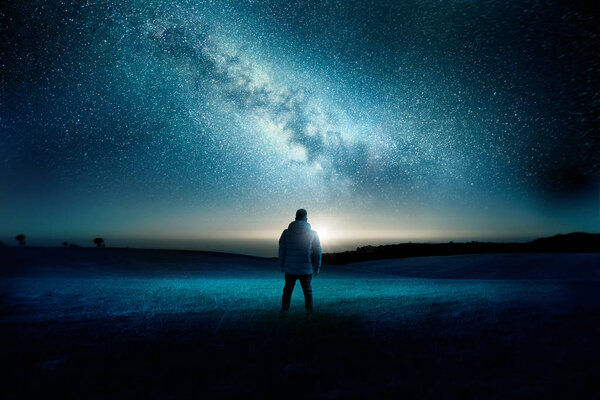 A man stands watching with wonder and amazement as the moon and milky way galaxy fill the night sky. Night time landscape. Photo composite.