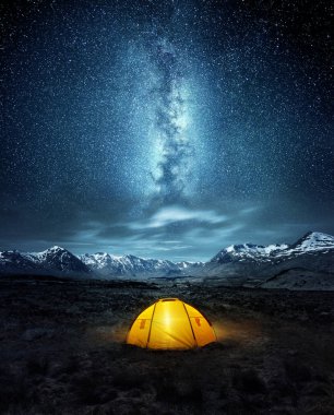 Camping in the wilderness. A pitched tent under the glowing  night sky stars of the milky way with snowy mountains in the background. Nature landscape photo composite. clipart