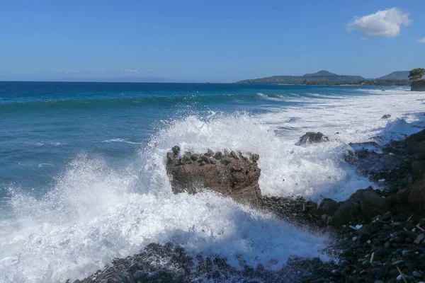 Le onde si infrangono su una spiaggia rocciosa, formando un grande spray. Grandi onde si infrangono contro le rocce durante una tempesta ai tropici. Potenti onde d'urto contro la roccia nera a Bali, Indonesia — Foto Stock