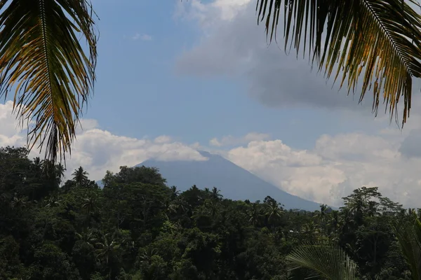 View through the trees in the foreground to the active Gunung Agung volcano from the opposite hill on Bali Island in Indonesia. Azure blue sky with dense white clouds on the sides of the mountain.