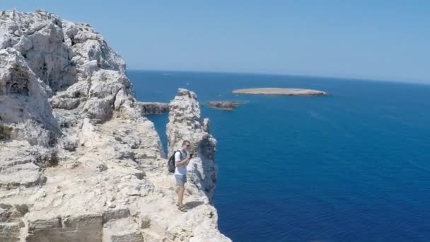 Lockdown-Aufnahme aus dem Rücken eines jungen Mannes, der Fotos vom Meer macht. Ein junger Mann fotografiert auf der Klippe des Cap Cavaleria, Menorca, Spanien. Sommerzeit in Spanien.