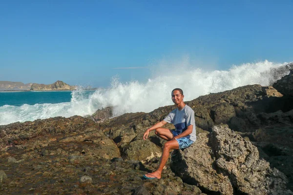 Un homme est assis sur une pierre près de l'océan. Des vagues se brisent sur une falaise rocheuse. Adolescent près de la mer orageuse. Jeune homme sur une falaise avec vue sur la mer. Un jeune Asiatique en short et un T-shirt assis sur une falaise — Photo