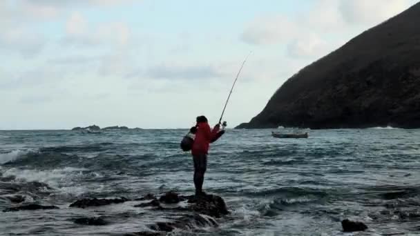 Pescador con ropa negra sostiene caña de pescar en sus manos. Un pescador pescó un pez en un arrecife en la marea alta. Un hombre con una caña de pescar atrapa peces en el océano — Vídeos de Stock