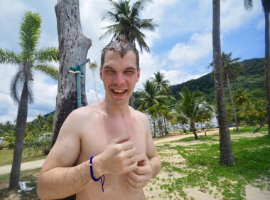 Sakin tropikal okyanus ve ağaçlar yakınındaki kumlu bir plajda bir palmiye ağacından asılı bir duş alarak gülümseyen genç sevimli fitness adam üstsüz renkli portre