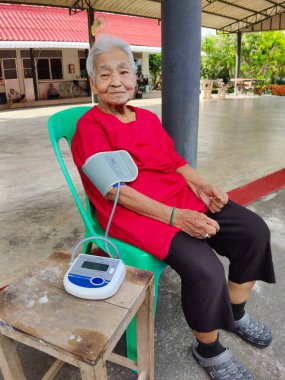 CHIANG RAI, THAILAND - 7 Haziran 2020 'de Tayland, Chiang Rai' de tansiyon monitörü kullanarak yeşil sandalyede oturan kimliği belirsiz yaşlı kadın..