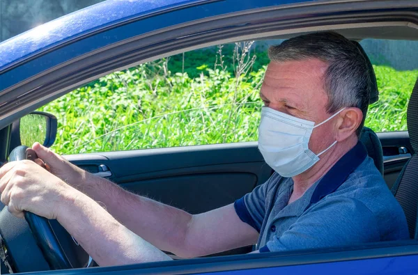 Orta yaşlı bir adam özel bir arabanın direksiyonunda oturuyor tıbbi maske takıyor ve önüne bakıyor. Salgın covid-19, kendini tecrit, maske konsepti. Yatay yönelim, seçici odak.