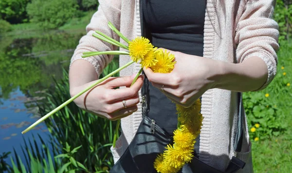 Woman hands weaving  wreath of dandelions on bank of  beautiful lake or river