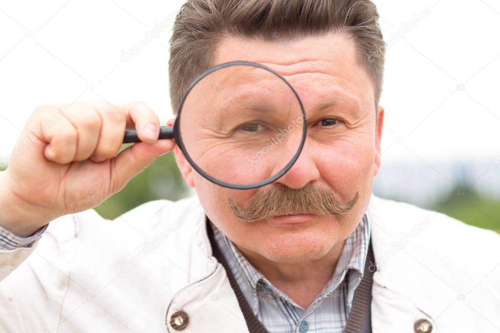 Close up face. Adult middle aged man with a mustache looks through a magnifying glass.