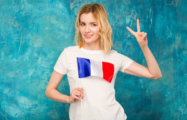 Beautiful blond woman with a flag of France in hands standing on a blue background. Show two fingers as a sign of luck or victory.