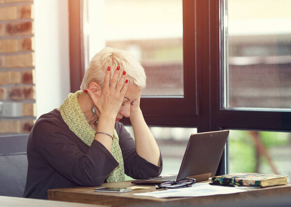 Adult woman thinking about to complete work task. Tired woman working at office desk in front of laptop suffering from chronic daily headaches from computer. Frustrated woman.