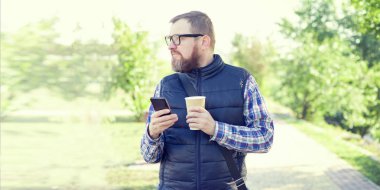 Adam bir cep telefonu ile ve bir fincan kahve sokakta yürürken. Gözlük ve sakallı bir adam.