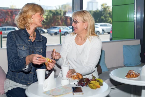 Two mature women drink coffee in a cafe. Seniors women longtime friends in travel have lunch at a roadside restaurant.