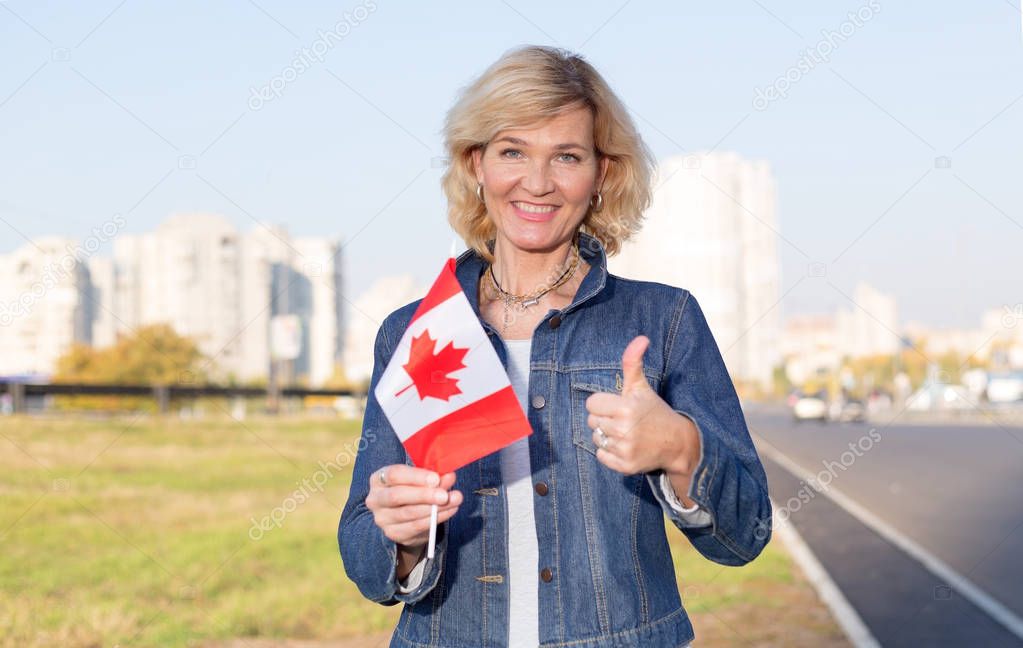 Mature woman with canada flag showing thumb up standing against city and blue sky.